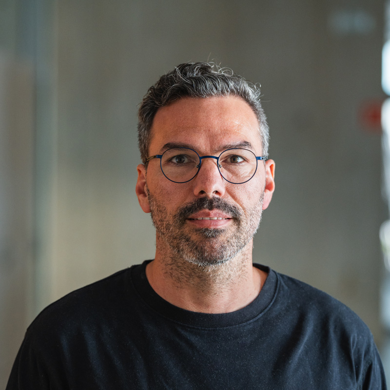 Portrait von Gregor Burger: graumelierte kurze Haare, Vollbart, runde Brille, schwarzes T-Shirt, vor unscharfem hellem Industrie-Hintergrund.