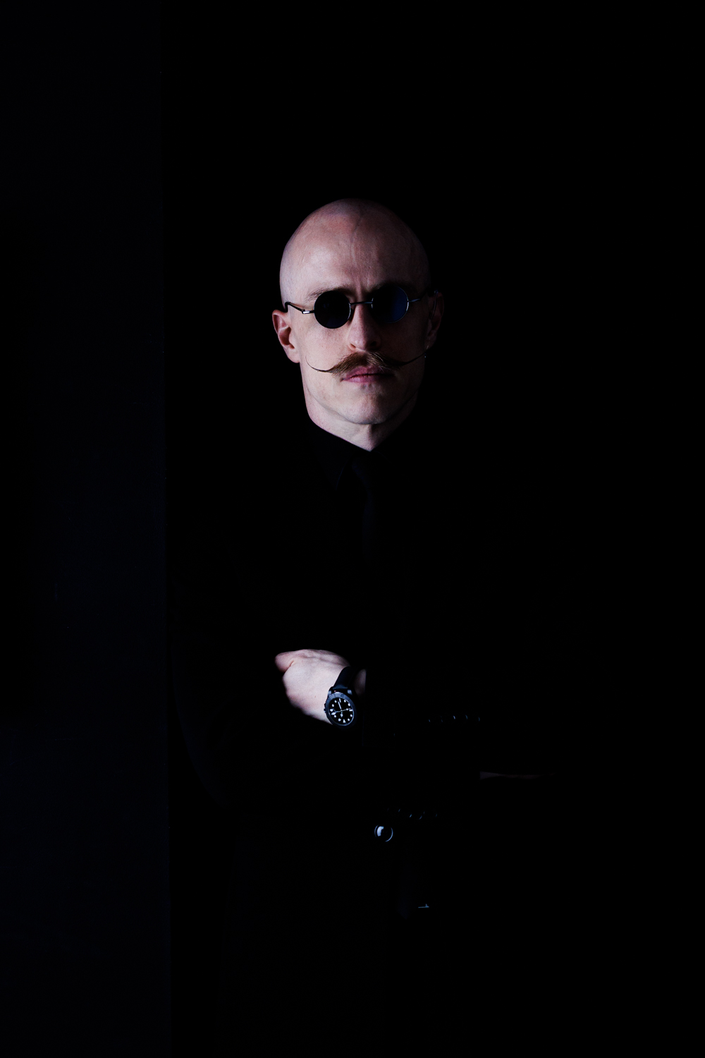 Portrait von Michael Mayer vor schwarzem Hintergrund: kahlrasiert, runde dunkle Sonnenbrille, gezwirbelter Schnurrbart, schwarzes Hemd, Arme verschränkt.