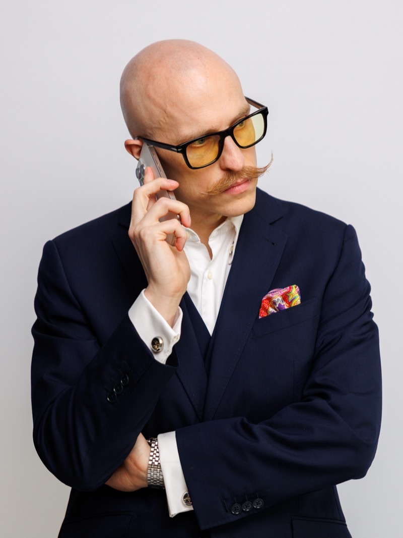 Michael Mayer, Gründer von nullzwo, am Telefon: dunkelblauer Anzug, weißes Hemd, buntes Einstecktuch, gelb getönte Brille, gezwirbelter Schnurrbart, Smartphone am Ohr, Blick zur Seite.